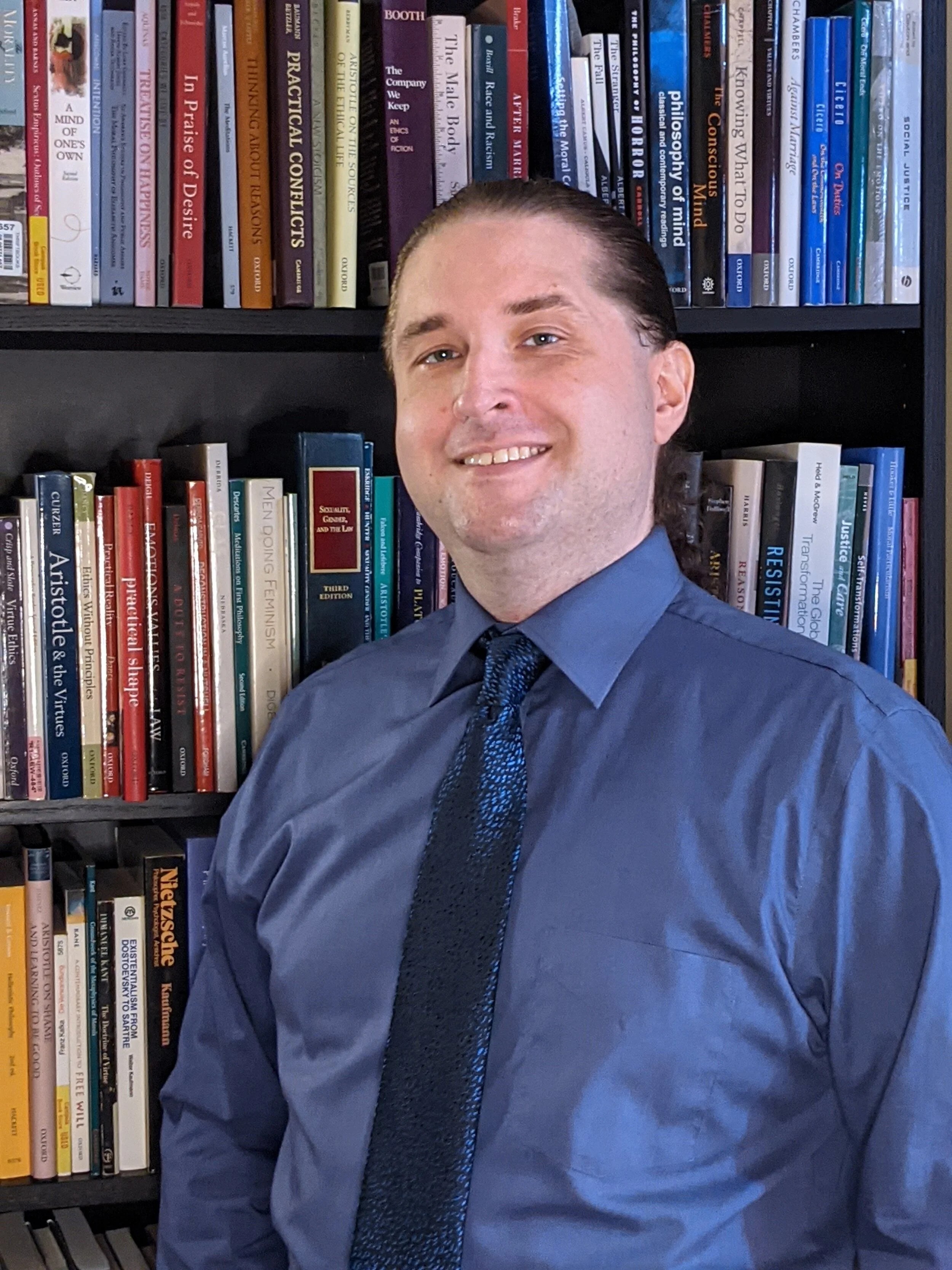 Duane Long in a blue shirt and tie smiling in front of a bookcase